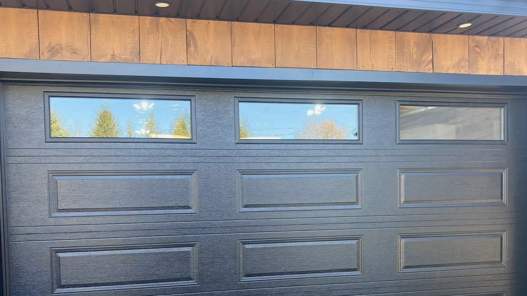 Porte de garage noire à fenêtres, sous un revêtement de bois